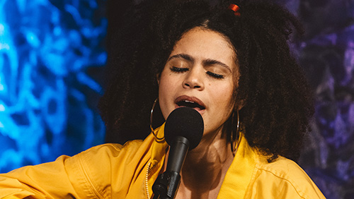 Arts collections. Black woman in yellow jacket sings at a mic on stage