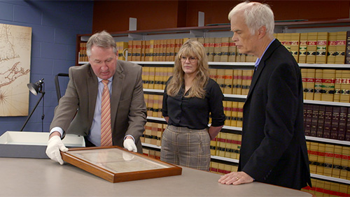 photo of two men and a woman in a library examining an artifact in a frame