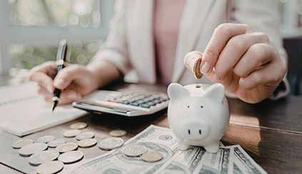 Photo of a woman putting a coin into a piggy bank with money, a notebook, and a calculator on a desk